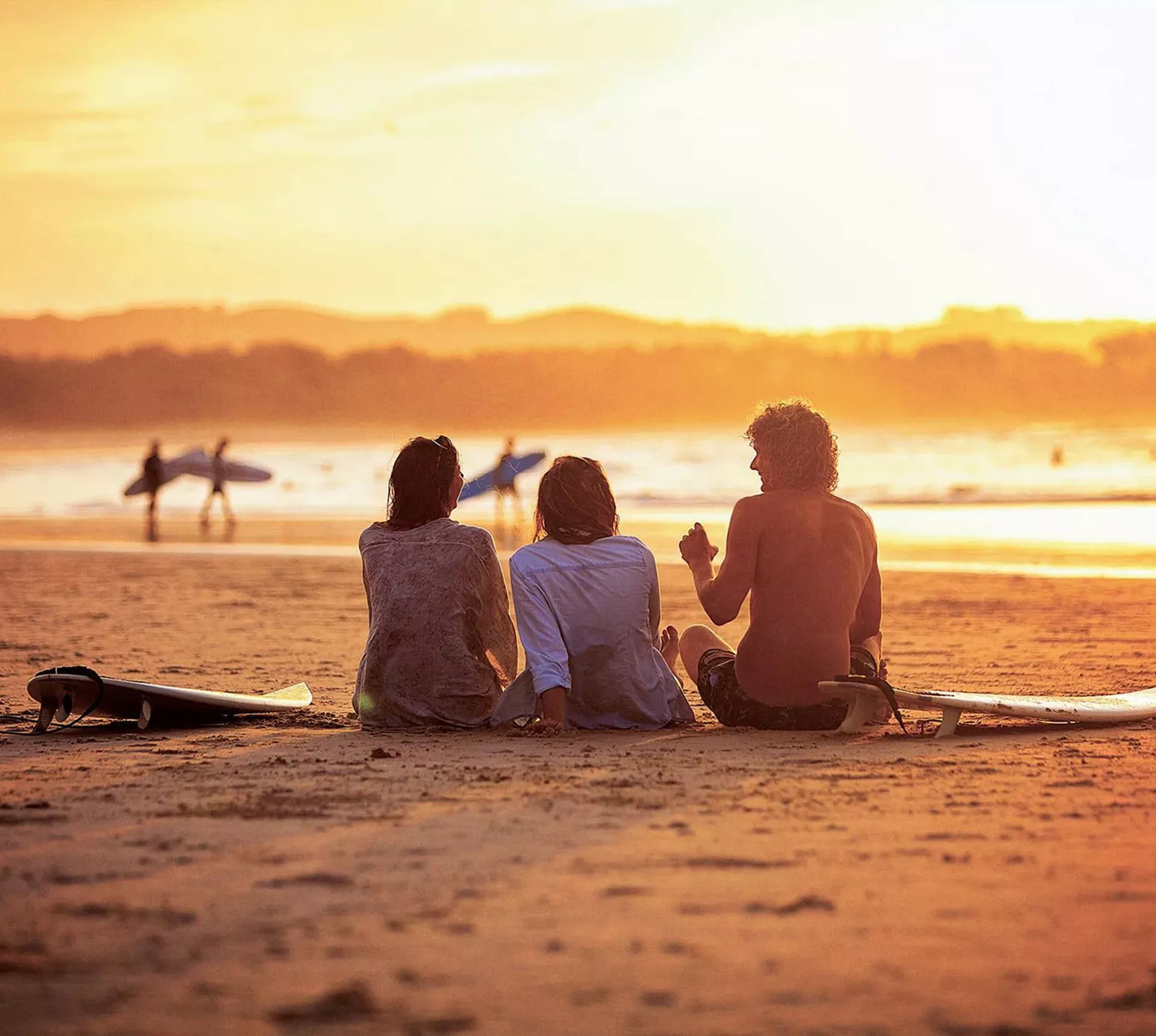 Contiki travellers relaxing on the sand with surfboards watching surfers walk along the shoreline at a stunning golden sunset in Byron Bay Australia