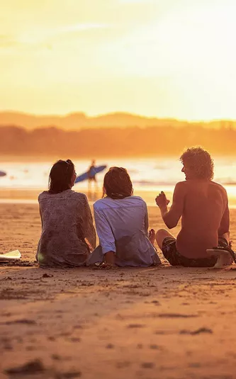 Contiki travellers relaxing on the sand with surfboards watching surfers walk along the shoreline at a stunning golden sunset in Byron Bay Australia