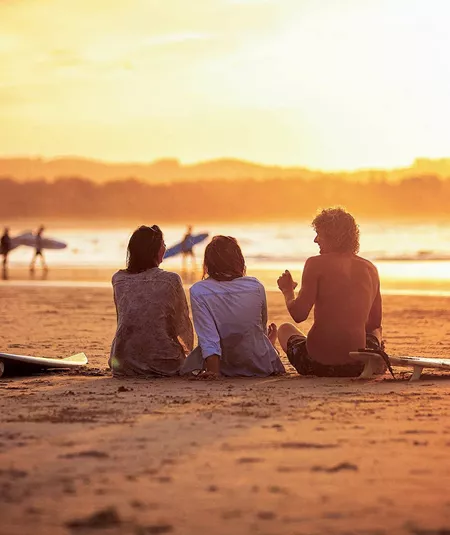 Contiki travellers relaxing on the sand with surfboards watching surfers walk along the shoreline at a stunning golden sunset in Byron Bay Australia
