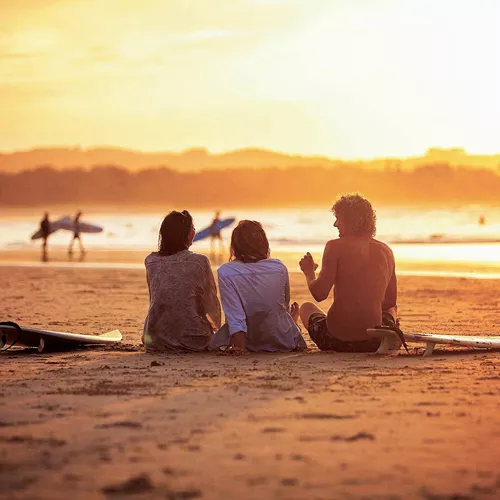 Contiki travellers relaxing on the sand with surfboards watching surfers walk along the shoreline at a stunning golden sunset in Byron Bay Australia