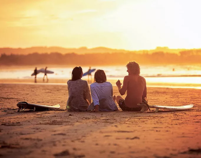 Contiki travellers relaxing on the sand with surfboards watching surfers walk along the shoreline at a stunning golden sunset in Byron Bay Australia