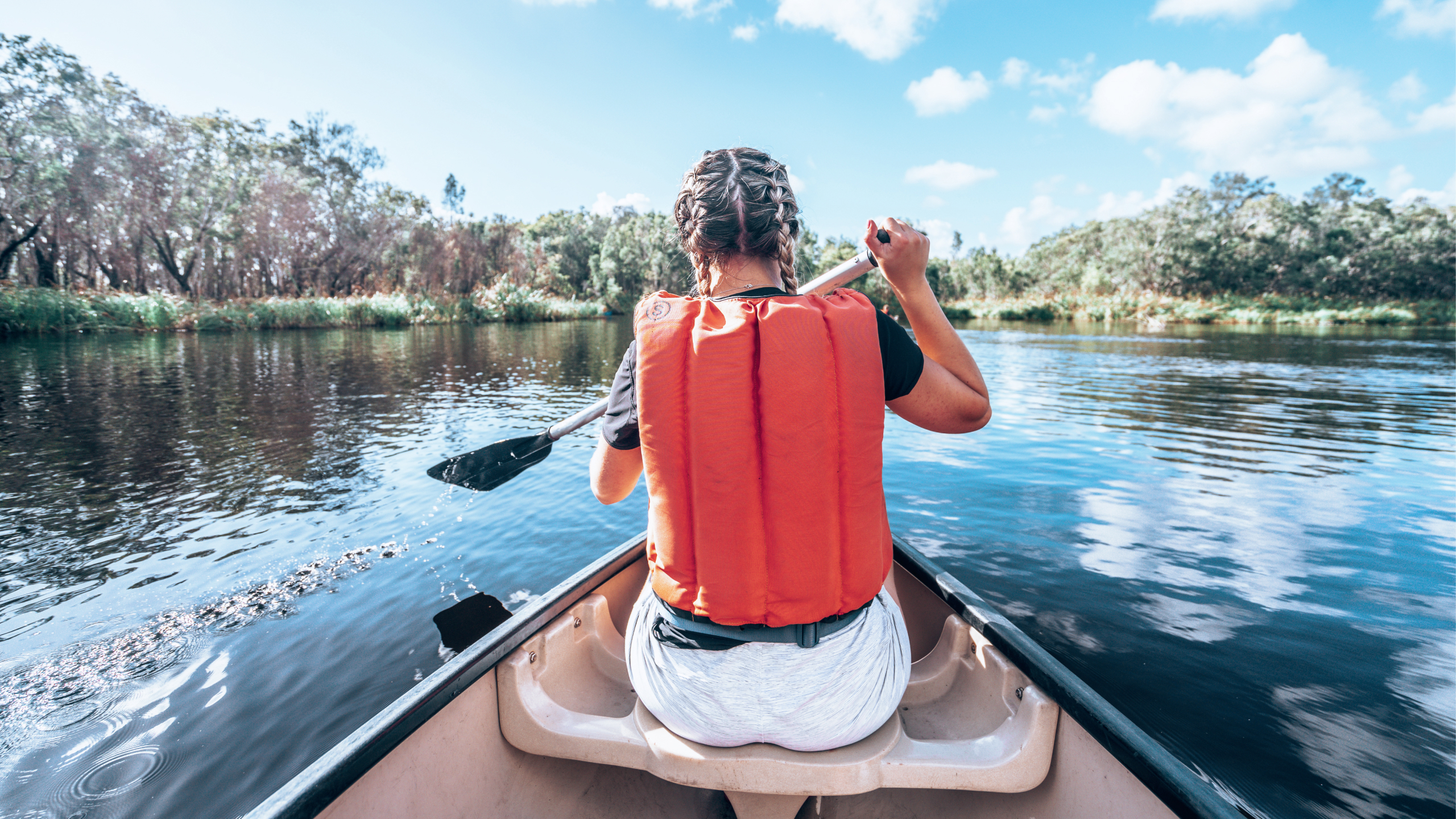 A Contiki travellers paddles a canoe across calm waters, surrounded by native bushland and mirrored reflections in the peaceful wetlands - Sydney to Noosa Tour