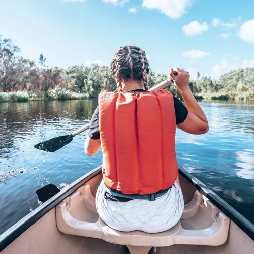 A Contiki travellers paddles a canoe across calm waters, surrounded by native bushland and mirrored reflections in the peaceful wetlands - Sydney to Noosa Tour