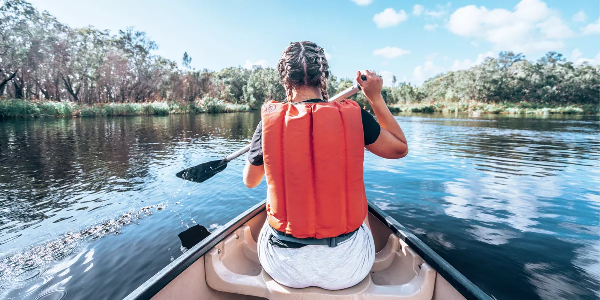 A Contiki travellers paddles a canoe across calm waters, surrounded by native bushland and mirrored reflections in the peaceful wetlands - Sydney to Noosa Tour