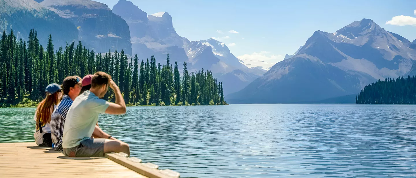 People Sitting Close To A Lake Enjoying The Snowy Mountains View