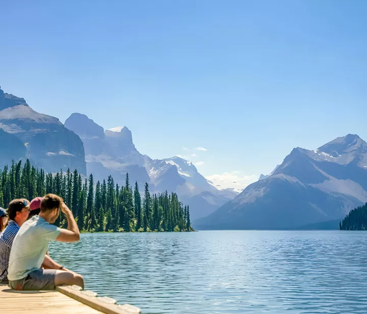 People Sitting Close To A Lake Enjoying The Snowy Mountains View