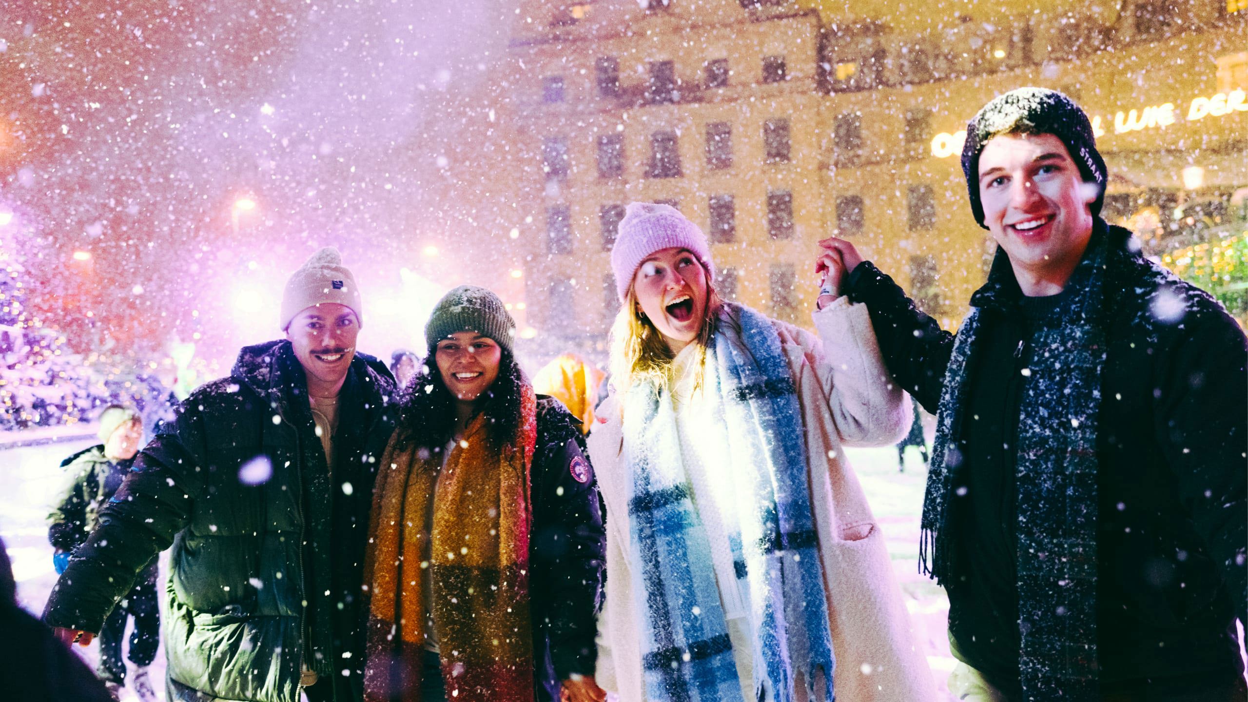 Group of four young travellers laughing in the snow at a festive Christmas market at night - Contiki German Christmas Markets Tour