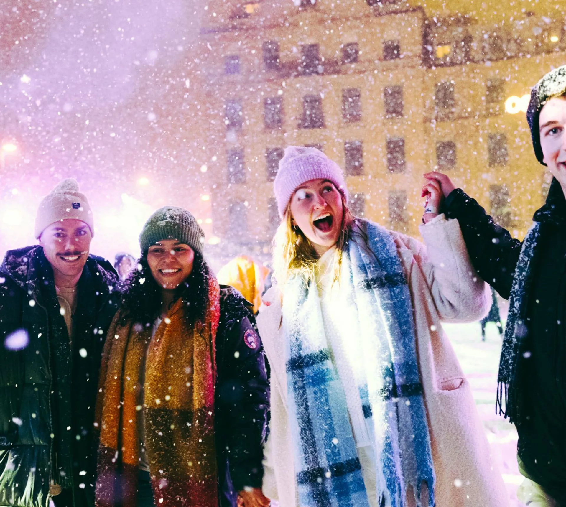Group of four young travellers laughing in the snow at a festive Christmas market at night - Contiki German Christmas Markets Tour
