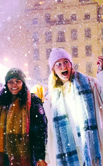 Group of four young travellers laughing in the snow at a festive Christmas market at night - Contiki German Christmas Markets Tour