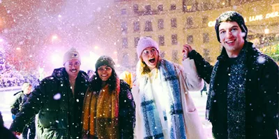 Group of four young travellers laughing in the snow at a festive Christmas market at night - Contiki German Christmas Markets Tour