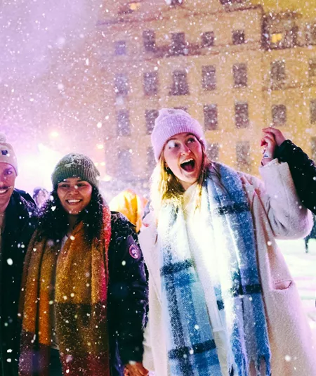Group of four young travellers laughing in the snow at a festive Christmas market at night - Contiki German Christmas Markets Tour