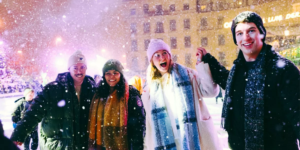Group of four young travellers laughing in the snow at a festive Christmas market at night - Contiki German Christmas Markets Tour