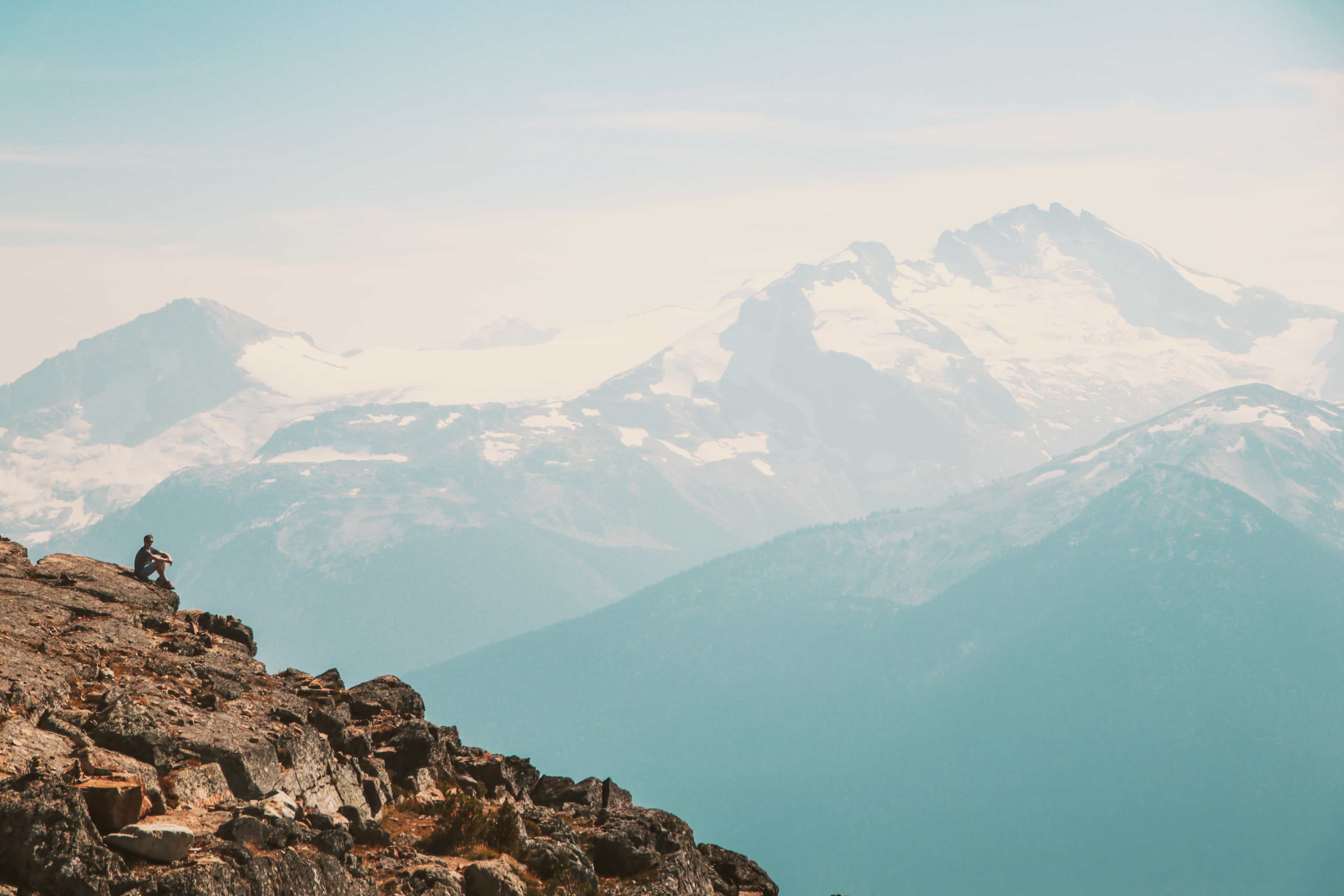Traveller sitting on a rocky ridge overlooking layers of snow-capped mountains, Whistler, Canada.
