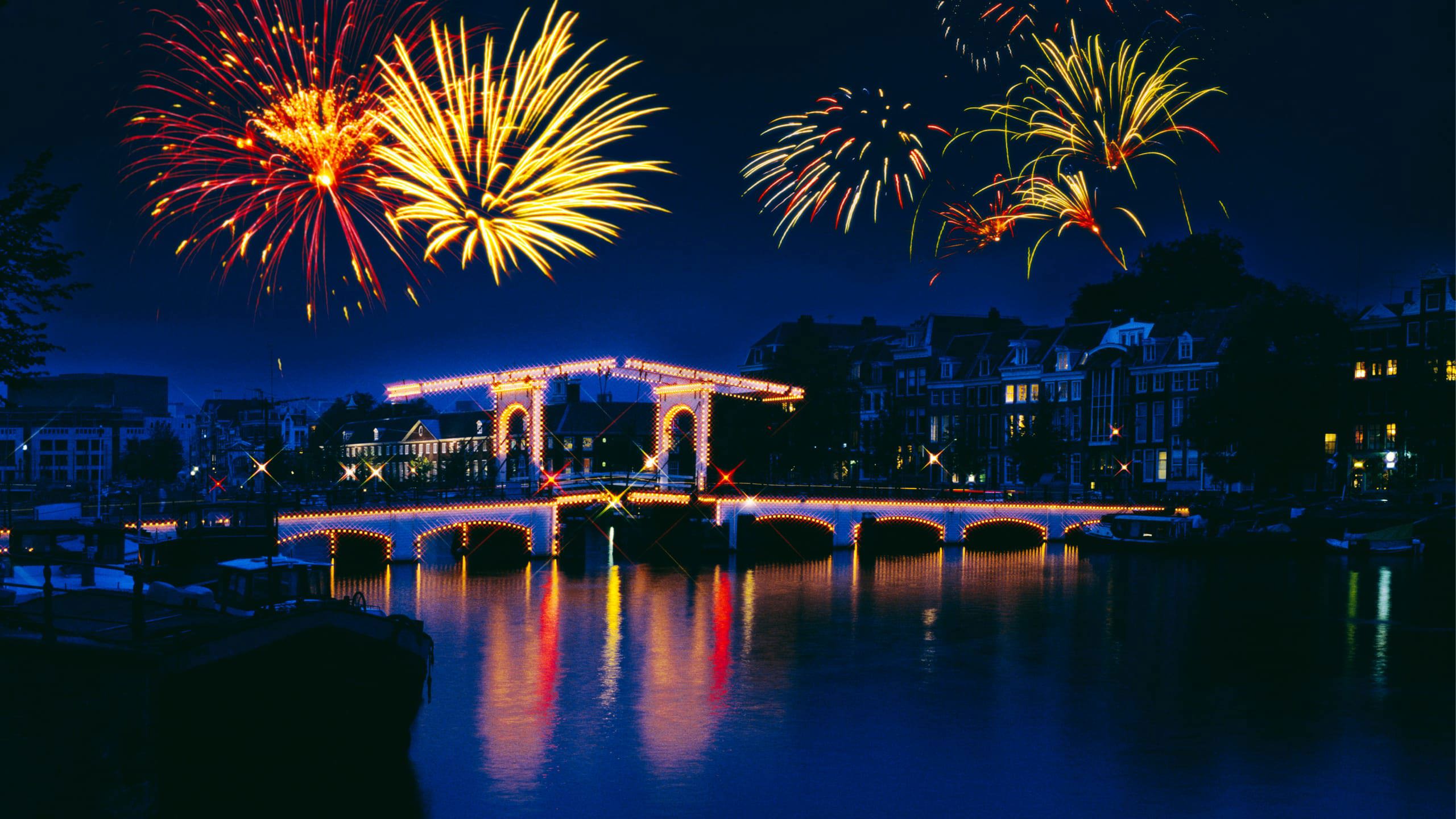 New Year's Eve fireworks lighting up the night sky over the illuminated Magere Brug bridge reflected in the Amstel River in Amsterdam Netherlands - Contiki Amsterdam New Year's Eve Tour