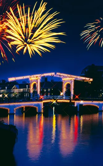 New Year's Eve fireworks lighting up the night sky over the illuminated Magere Brug bridge reflected in the Amstel River in Amsterdam Netherlands - Contiki Amsterdam New Year's Eve Tour