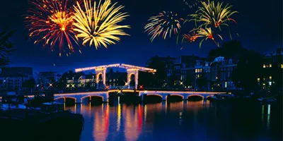 New Year's Eve fireworks lighting up the night sky over the illuminated Magere Brug bridge reflected in the Amstel River in Amsterdam Netherlands - Contiki Amsterdam New Year's Eve Tour
