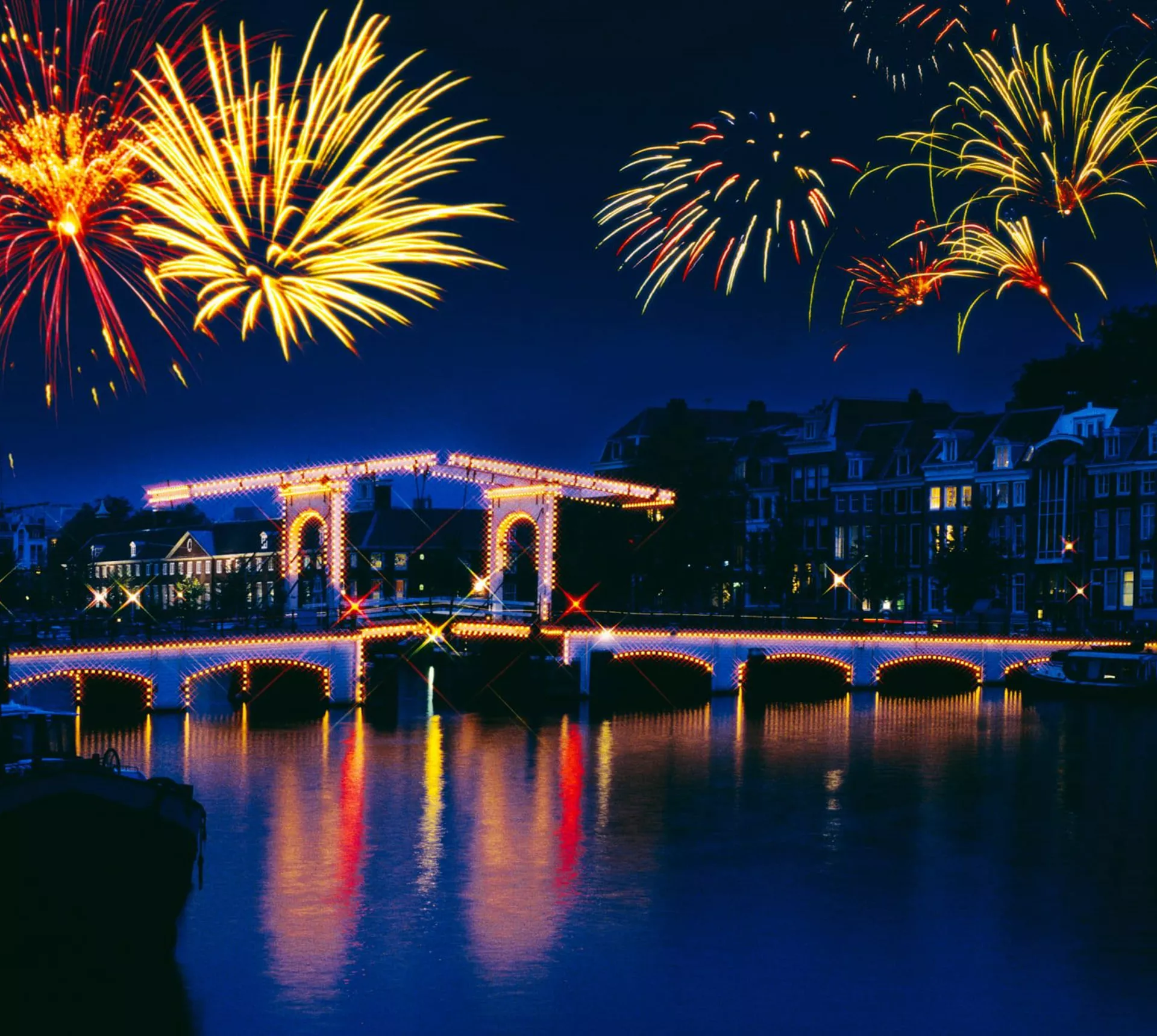 New Year's Eve fireworks lighting up the night sky over the illuminated Magere Brug bridge reflected in the Amstel River in Amsterdam Netherlands - Contiki Amsterdam New Year's Eve Tour
