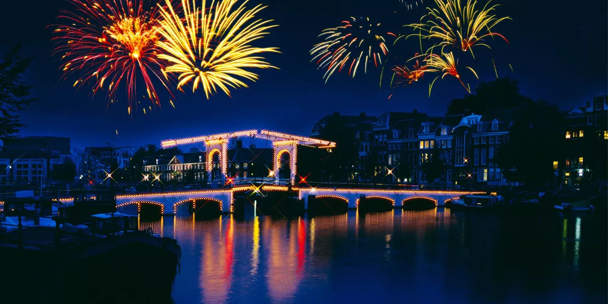 New Year's Eve fireworks lighting up the night sky over the illuminated Magere Brug bridge reflected in the Amstel River in Amsterdam Netherlands - Contiki Amsterdam New Year's Eve Tour
