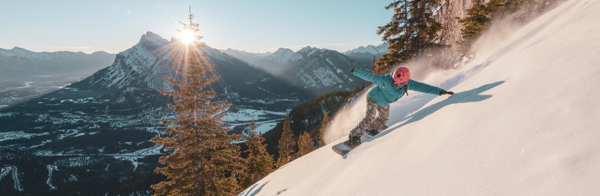 Snowboarder carving down a steep snowy slope with sunlit mountains in the background