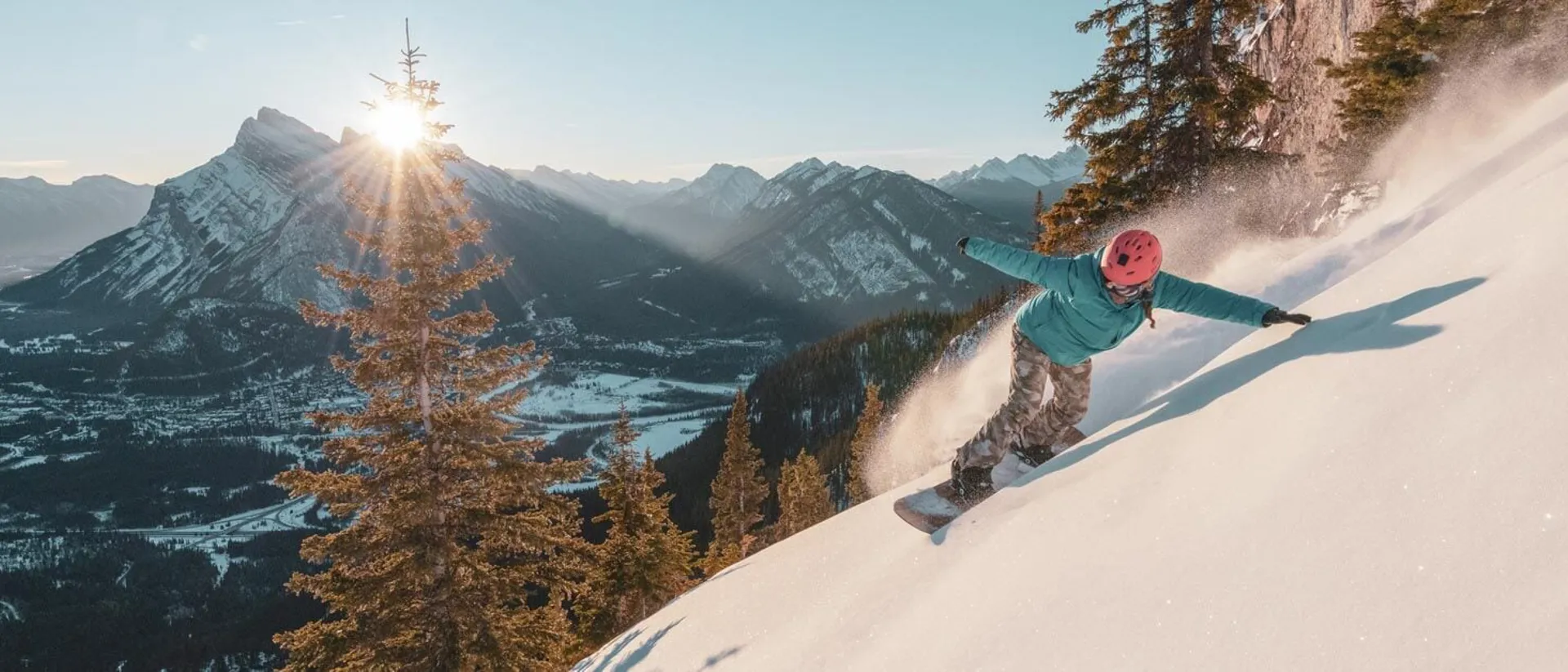 Snowboarder carving down a steep snowy slope with sunlit mountains in the background