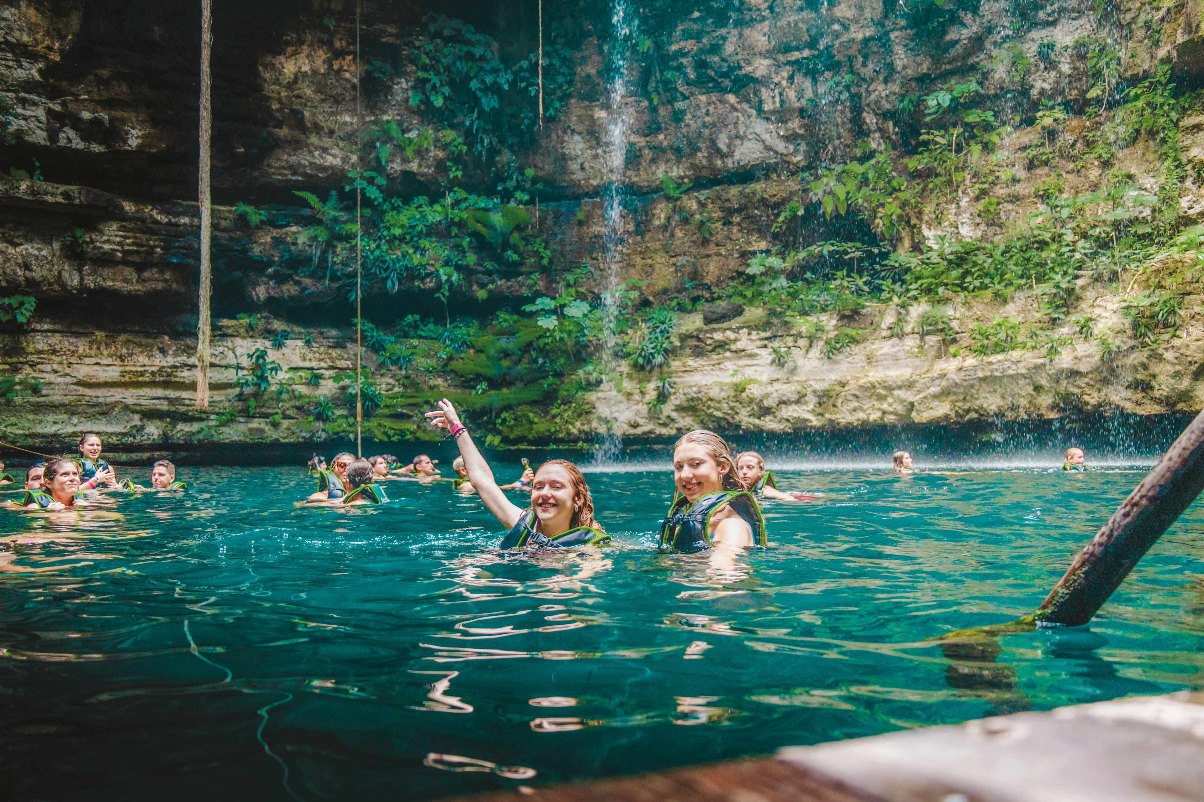 Two Women Swimming In A Natural Pool With A Group Of People