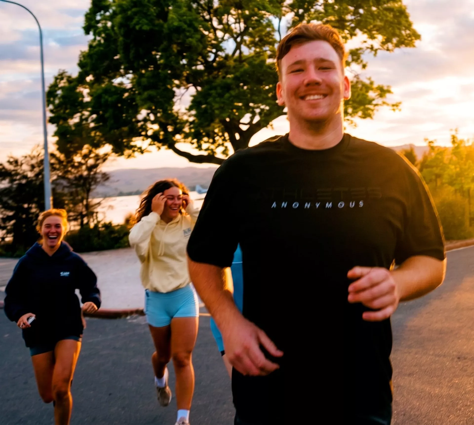 A group of travellers jog together along a lakeside road in New Zealand at sunset, smiling and enjoying the run - Contiki NZ Run Club: Muriwai Half Marathon Tour