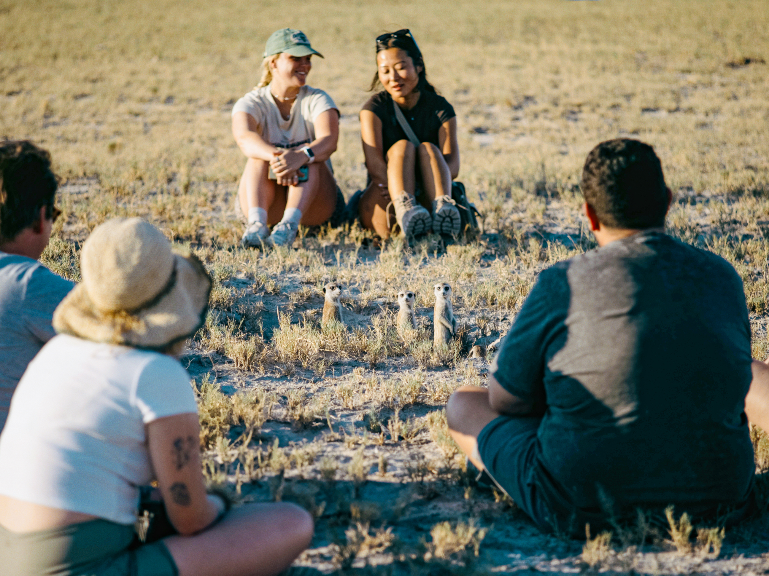 Travellers sitting down next to a meerkat in Makgadikgadi Salt Pans in Botswana, Africa