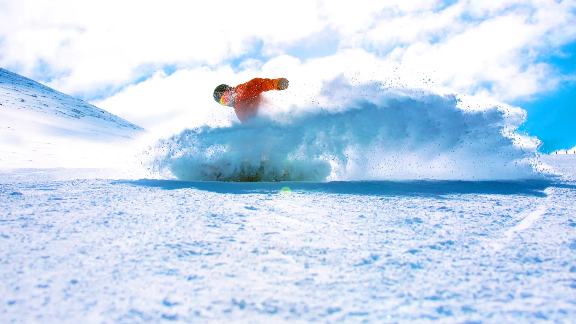 A snowboarder in a bright orange jacket rides through deep powder on a Canadian mountain, kicking up a spray of snow beneath a blue sky - Contiki Ski Canada Tour
