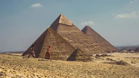 Two travellers walk across the sand beside the towering Pyramids of Giza.