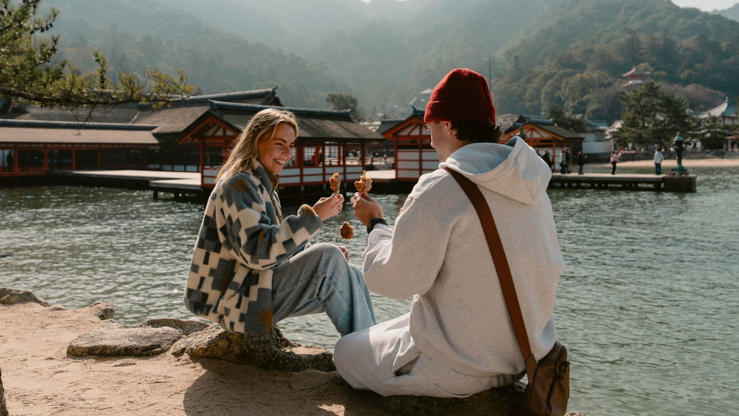 Two travellers enjoy grilled street food beside the waterfront on Miyajima Island, Japan, with the iconic Itsukushima Shrine and forested mountains in the background.