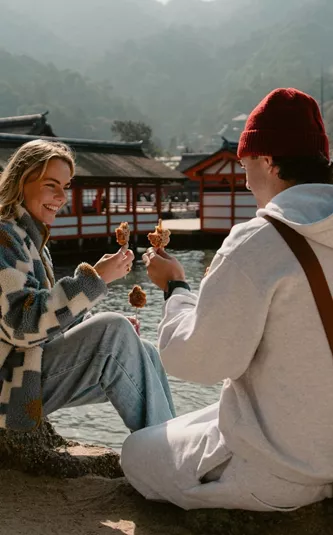 Two travellers enjoy grilled street food beside the waterfront on Miyajima Island, Japan, with the iconic Itsukushima Shrine and forested mountains in the background.