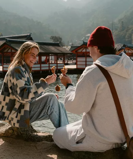 Two travellers enjoy grilled street food beside the waterfront on Miyajima Island, Japan, with the iconic Itsukushima Shrine and forested mountains in the background.