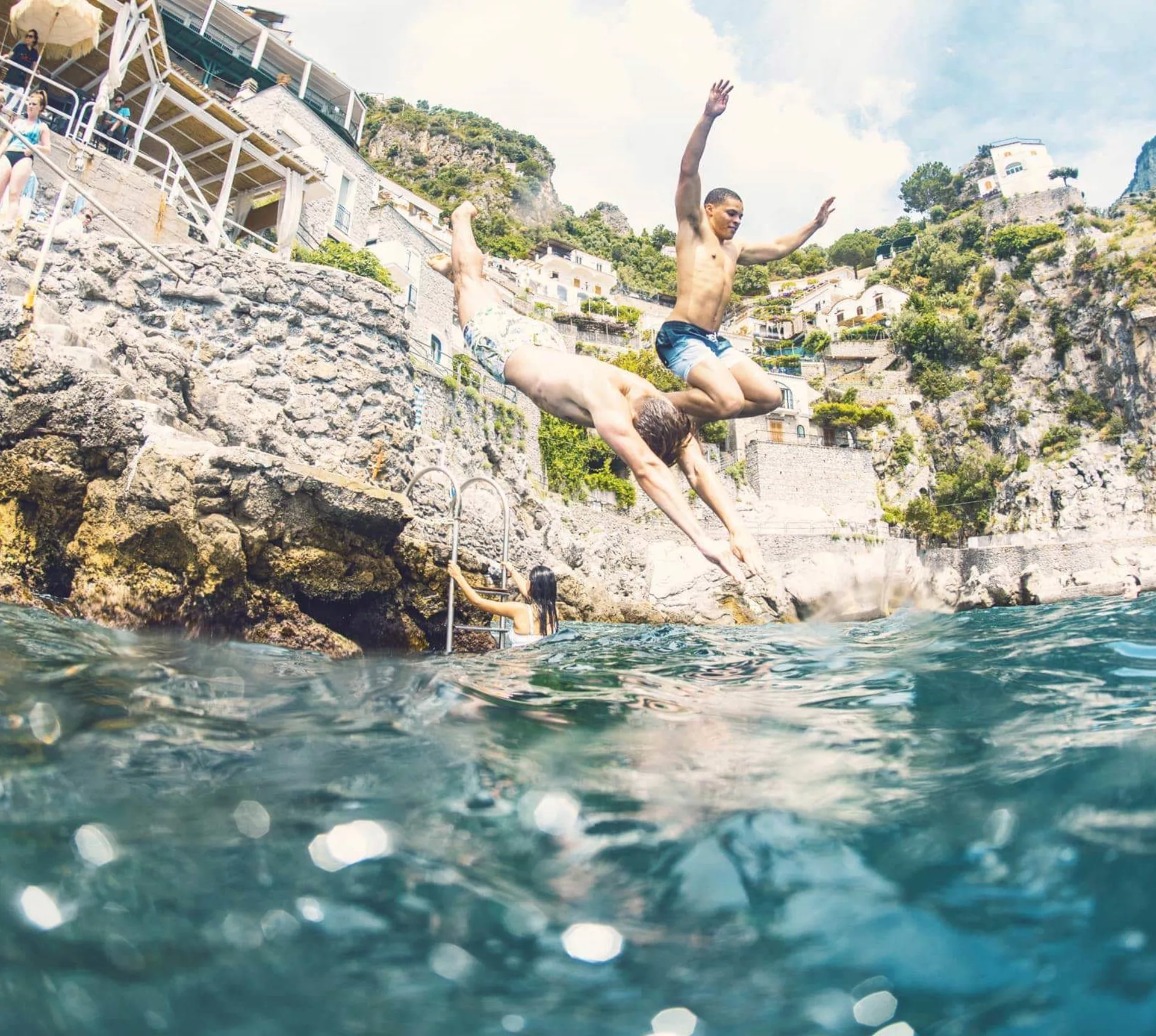 Young travellers jumping into the sea from rocks on the Amalfi Coast Italy with dramatic cliffside scenery - Contiki European Quest Tour