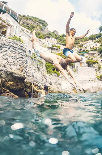 Young travellers jumping into the sea from rocks on the Amalfi Coast Italy with dramatic cliffside scenery - Contiki European Quest Tour