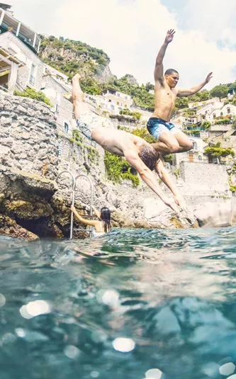 Young travellers jumping into the sea from rocks on the Amalfi Coast Italy with dramatic cliffside scenery - Contiki European Quest Tour