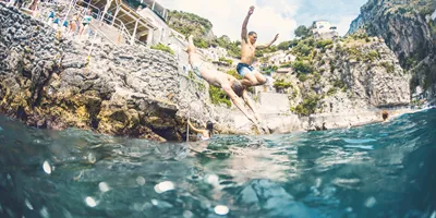 Young travellers jumping into the sea from rocks on the Amalfi Coast Italy with dramatic cliffside scenery - Contiki European Quest Tour