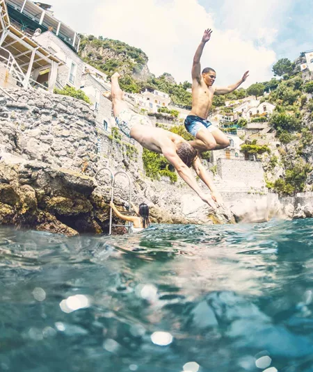 Young travellers jumping into the sea from rocks on the Amalfi Coast Italy with dramatic cliffside scenery - Contiki European Quest Tour