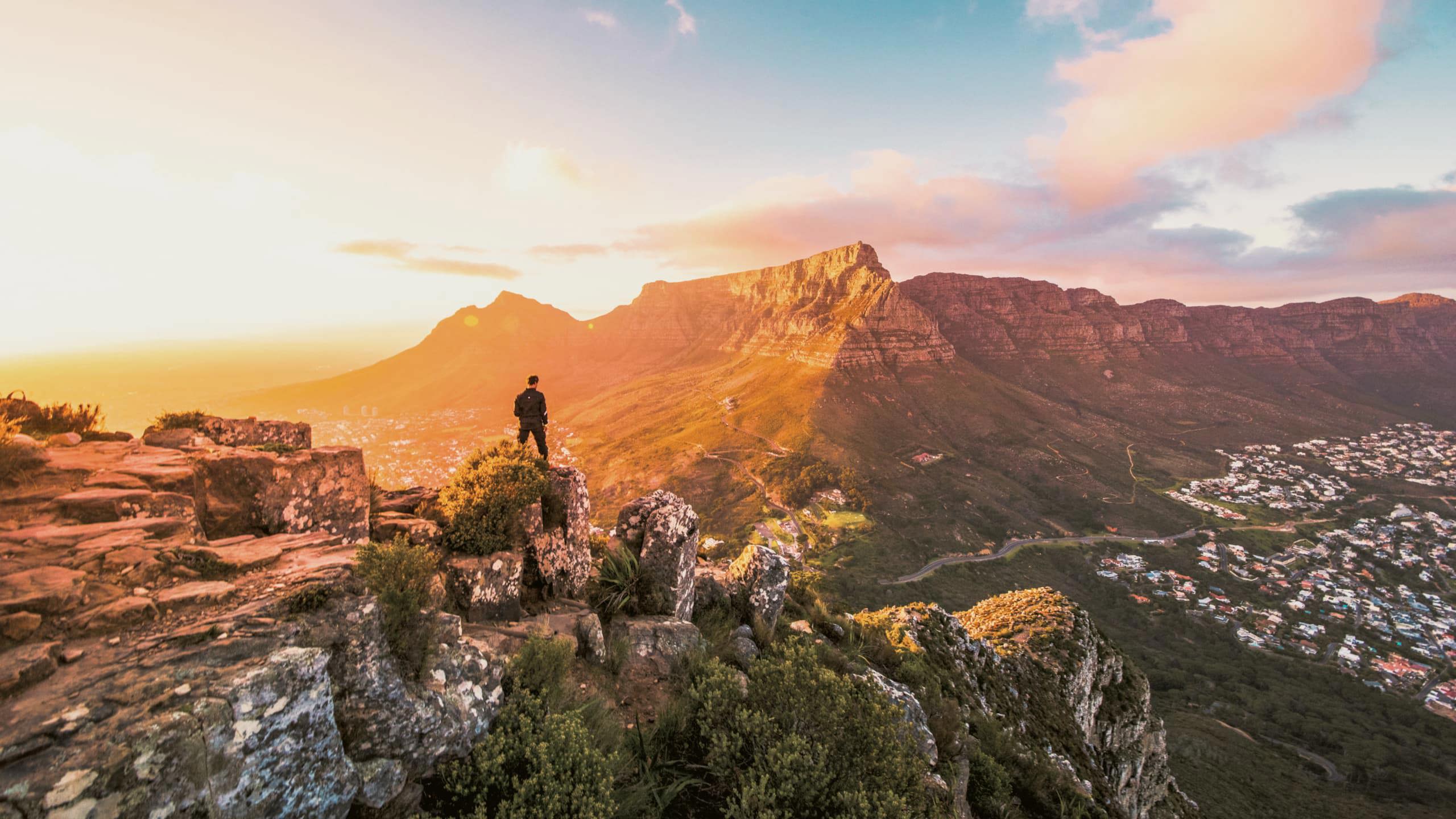 Solo traveller standing on rocky Lion's Head summit at golden hour overlooking the iconic Table Mountain and Cape Town cityscape in South Africa - Contiki Cape Town, Safari & Victoria Falls Tour