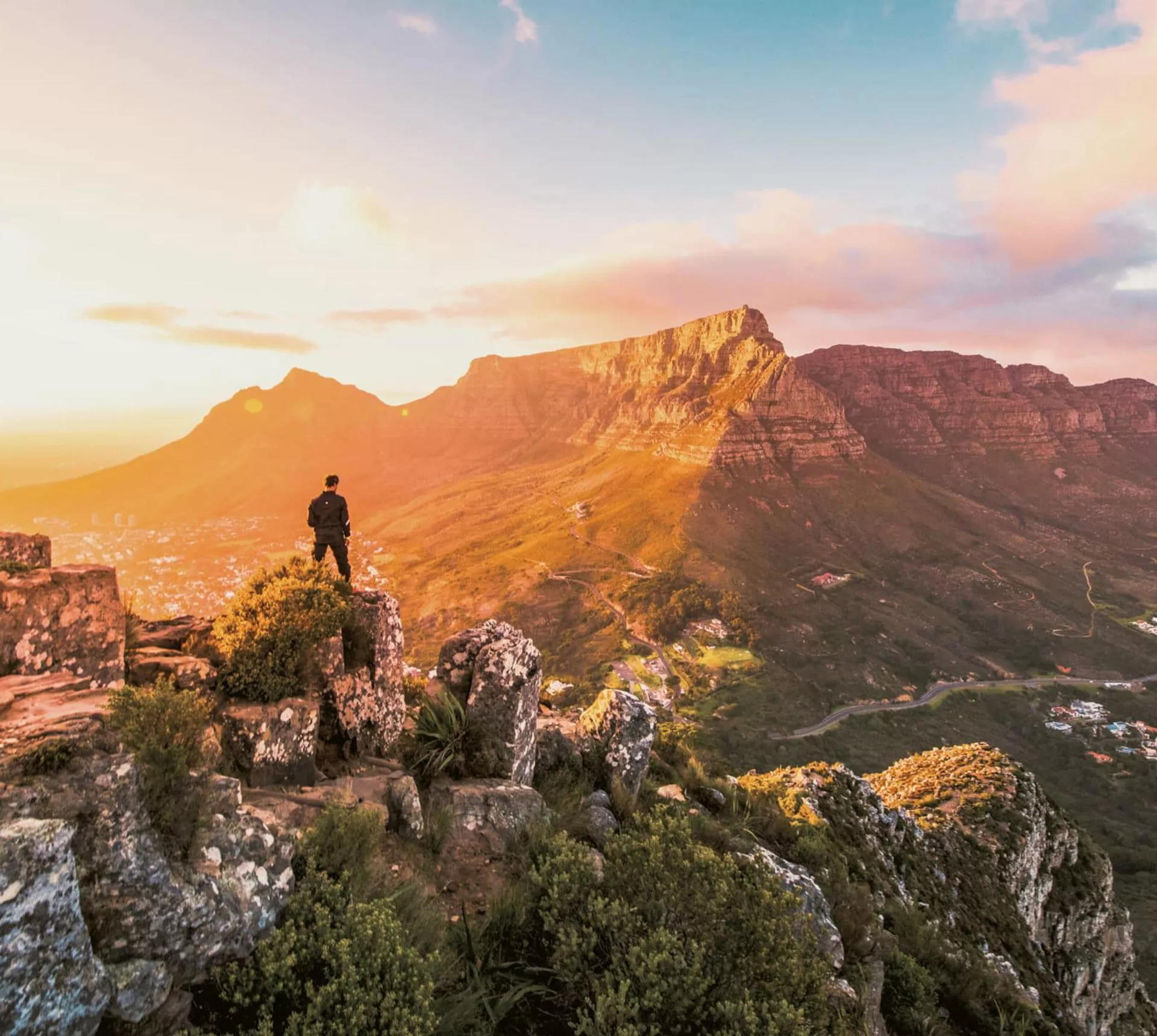Solo traveller standing on rocky Lion's Head summit at golden hour overlooking the iconic Table Mountain and Cape Town cityscape in South Africa - Contiki Cape Town, Safari & Victoria Falls Tour