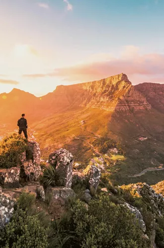 Solo traveller standing on rocky Lion's Head summit at golden hour overlooking the iconic Table Mountain and Cape Town cityscape in South Africa - Contiki Cape Town, Safari & Victoria Falls Tour