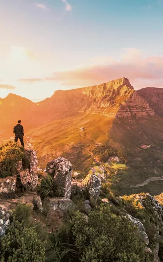 Solo traveller standing on rocky Lion's Head summit at golden hour overlooking the iconic Table Mountain and Cape Town cityscape in South Africa - Contiki Cape Town, Safari & Victoria Falls Tour