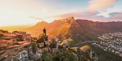 Solo traveller standing on rocky Lion's Head summit at golden hour overlooking the iconic Table Mountain and Cape Town cityscape in South Africa - Contiki Cape Town, Safari & Victoria Falls Tour