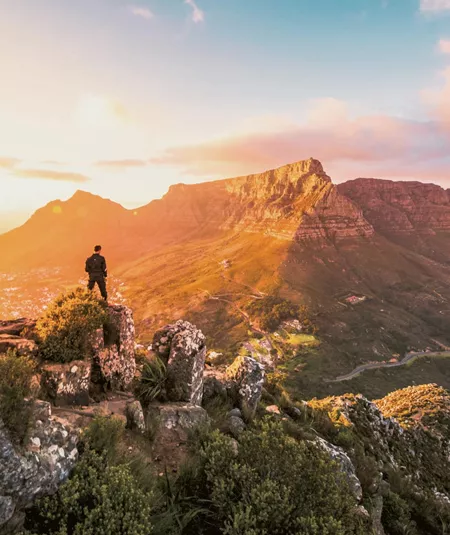 Solo traveller standing on rocky Lion's Head summit at golden hour overlooking the iconic Table Mountain and Cape Town cityscape in South Africa - Contiki Cape Town, Safari & Victoria Falls Tour