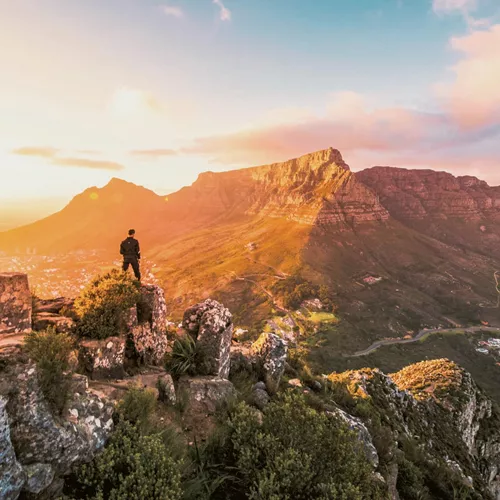 Solo traveller standing on rocky Lion's Head summit at golden hour overlooking the iconic Table Mountain and Cape Town cityscape in South Africa - Contiki Cape Town, Safari & Victoria Falls Tour