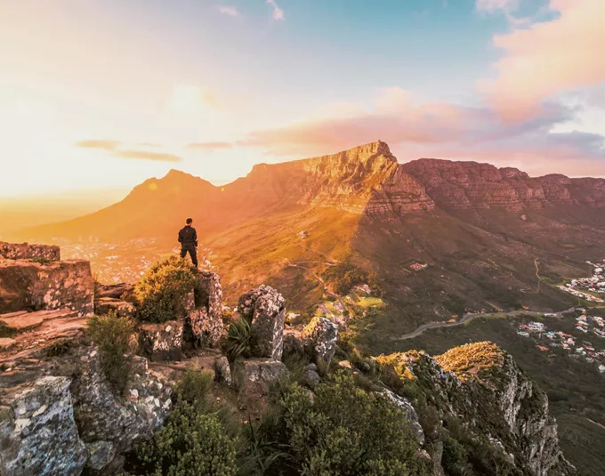 Solo traveller standing on rocky Lion's Head summit at golden hour overlooking the iconic Table Mountain and Cape Town cityscape in South Africa - Contiki Cape Town, Safari & Victoria Falls Tour