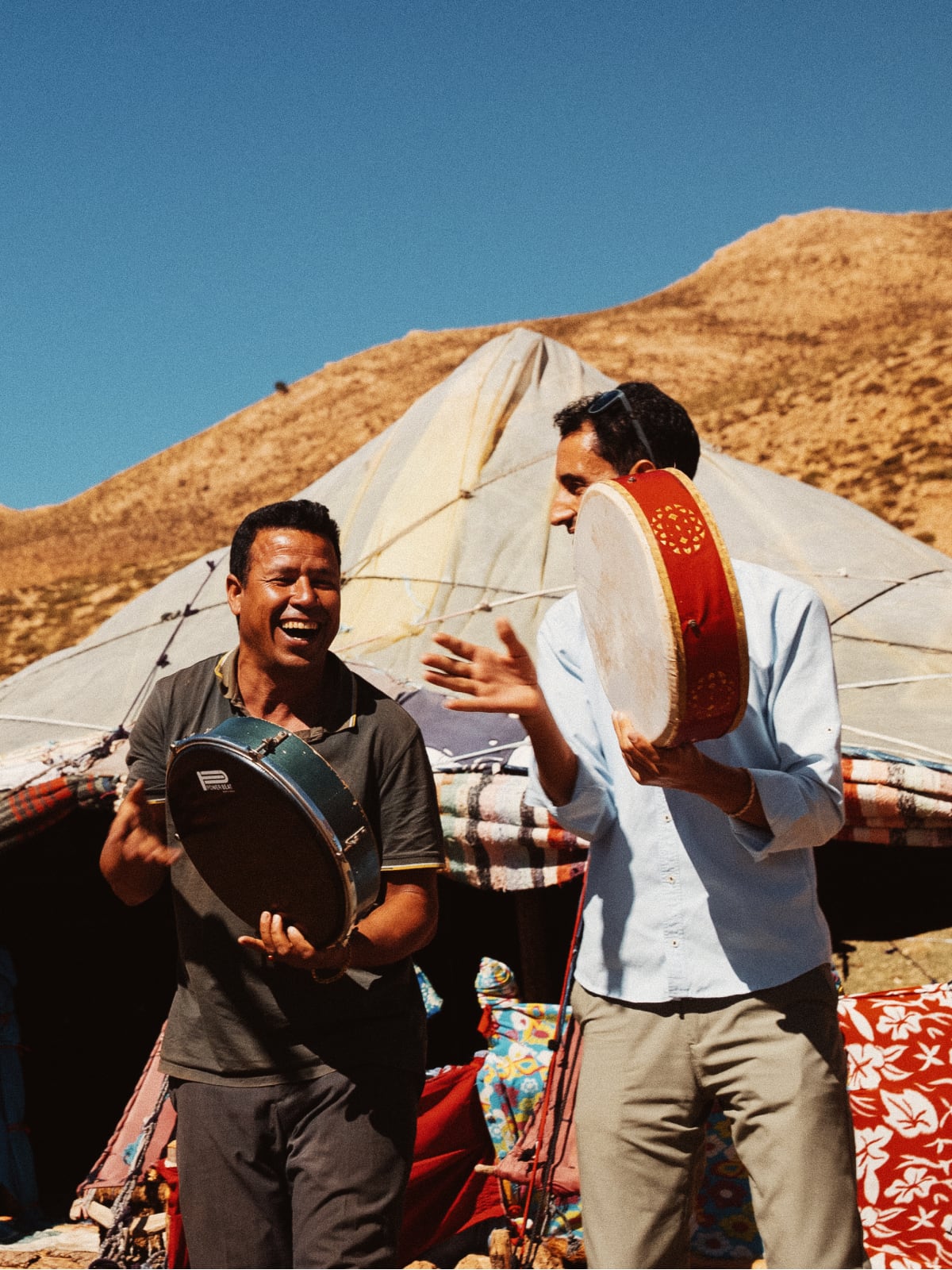 Two men laughing while playing traditional hand drums outside a Berber tent in the Moroccan desert.