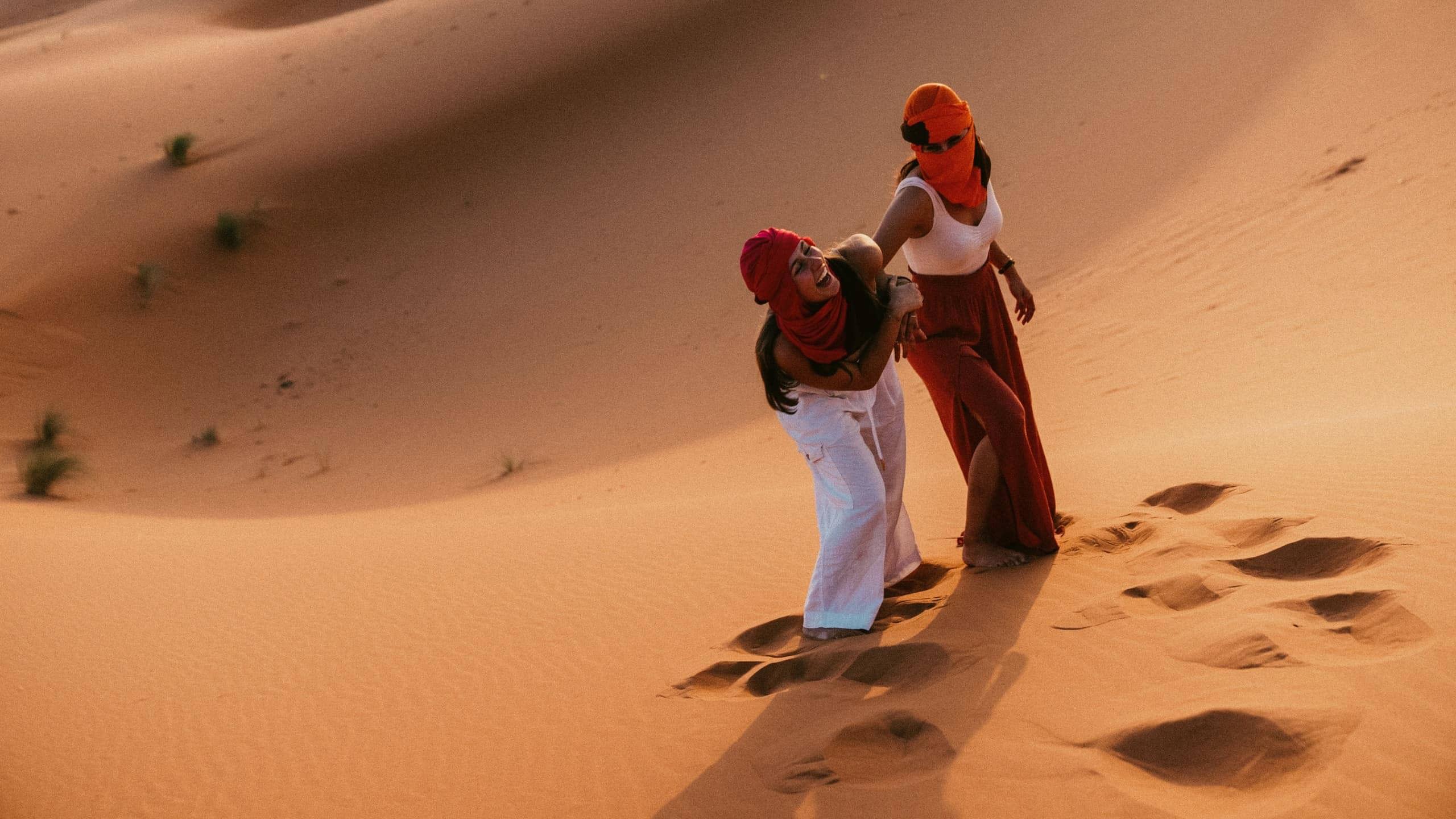 Two Contiki travellers climbing the golden sand dunes of the Sahara Desert in Morocco, dressed in traditional desert scarves during a sunset adventure on the Morocco Uncovered Tour