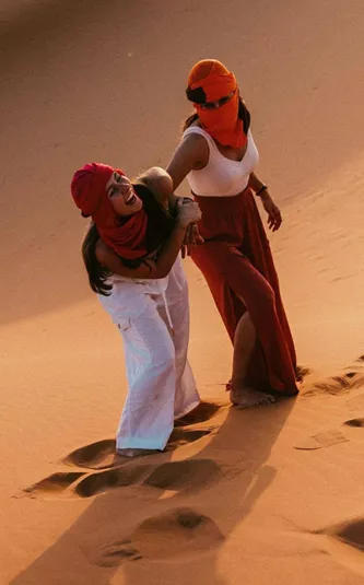 Two Contiki travellers climbing the golden sand dunes of the Sahara Desert in Morocco, dressed in traditional desert scarves during a sunset adventure on the Morocco Uncovered Tour