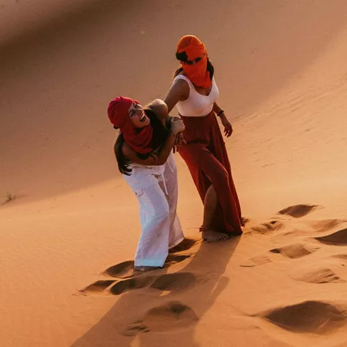 Two Contiki travellers climbing the golden sand dunes of the Sahara Desert in Morocco, dressed in traditional desert scarves during a sunset adventure on the Morocco Uncovered Tour