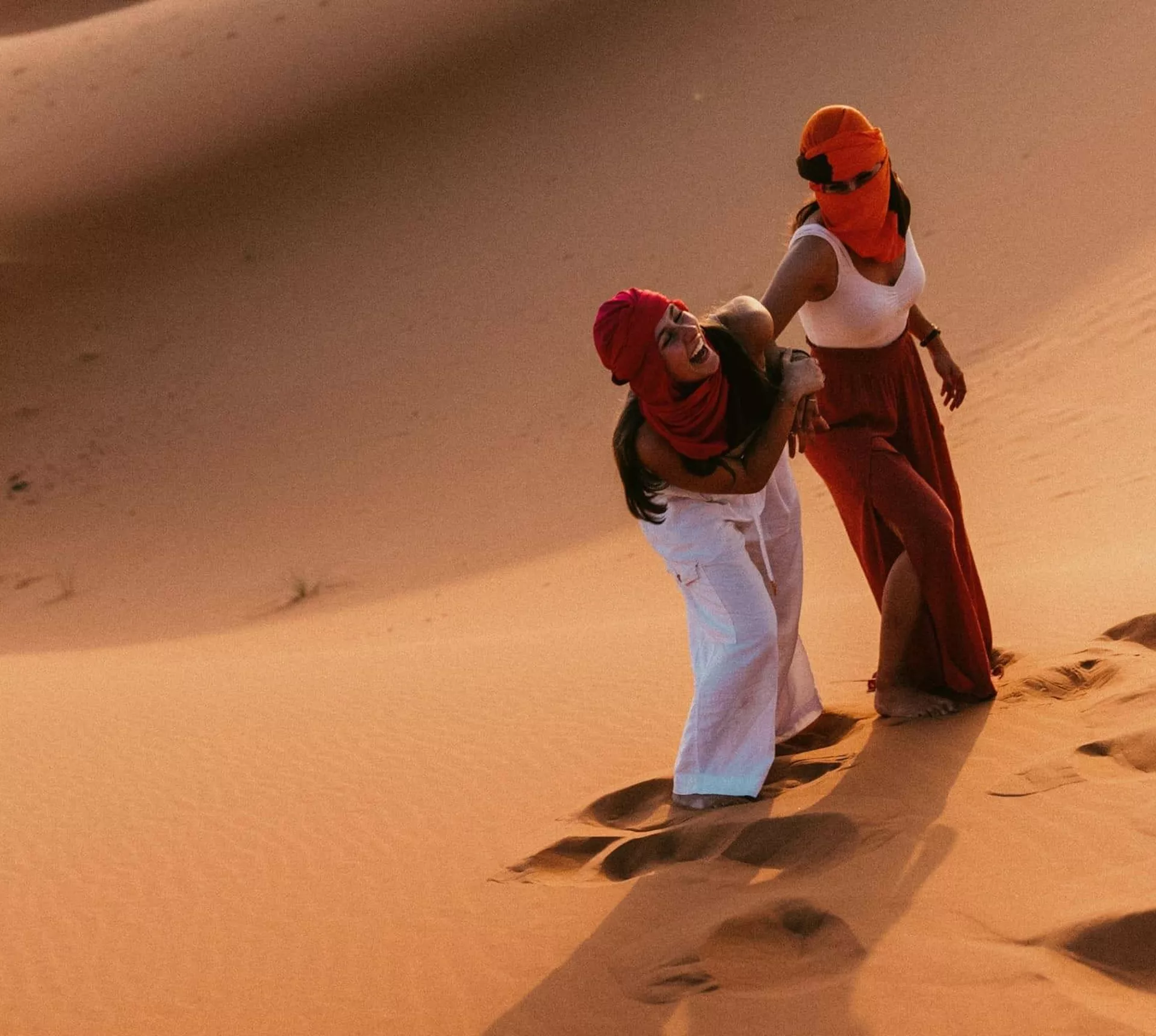 Two Contiki travellers climbing the golden sand dunes of the Sahara Desert in Morocco, dressed in traditional desert scarves during a sunset adventure on the Morocco Uncovered Tour
